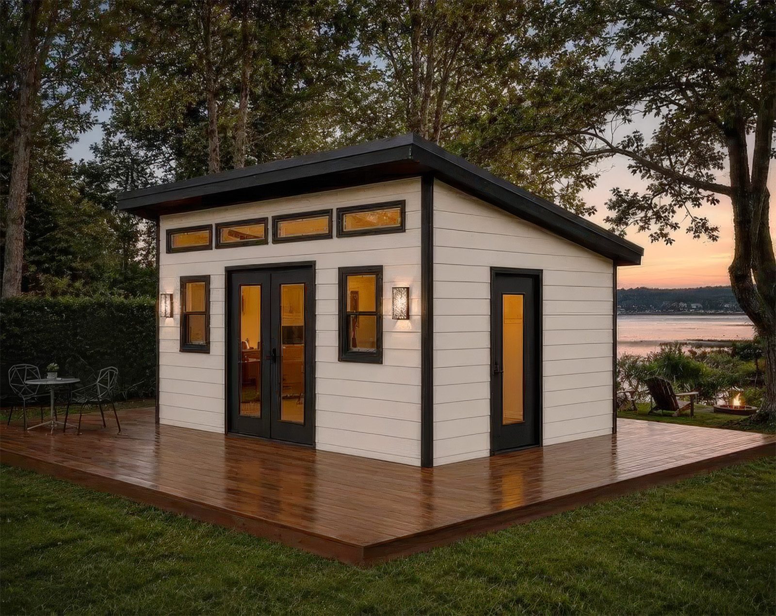 A modern backyard cottage at dusk: white lap siding, a sloped dark roof, clerestory windows, glass French doors and a wide wood deck on a lawn